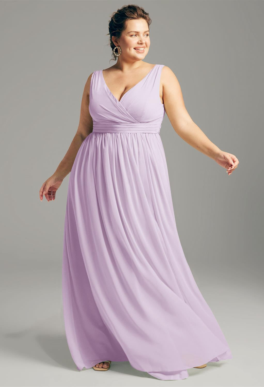 A woman poses against a neutral background wearing the Hilaria - Chiffon Bridesmaid Dress from Bergamot Bridal, a light purple sleeveless gown that embodies elegance, ideal for bridesmaids and available at bridal shops in London, Ontario.