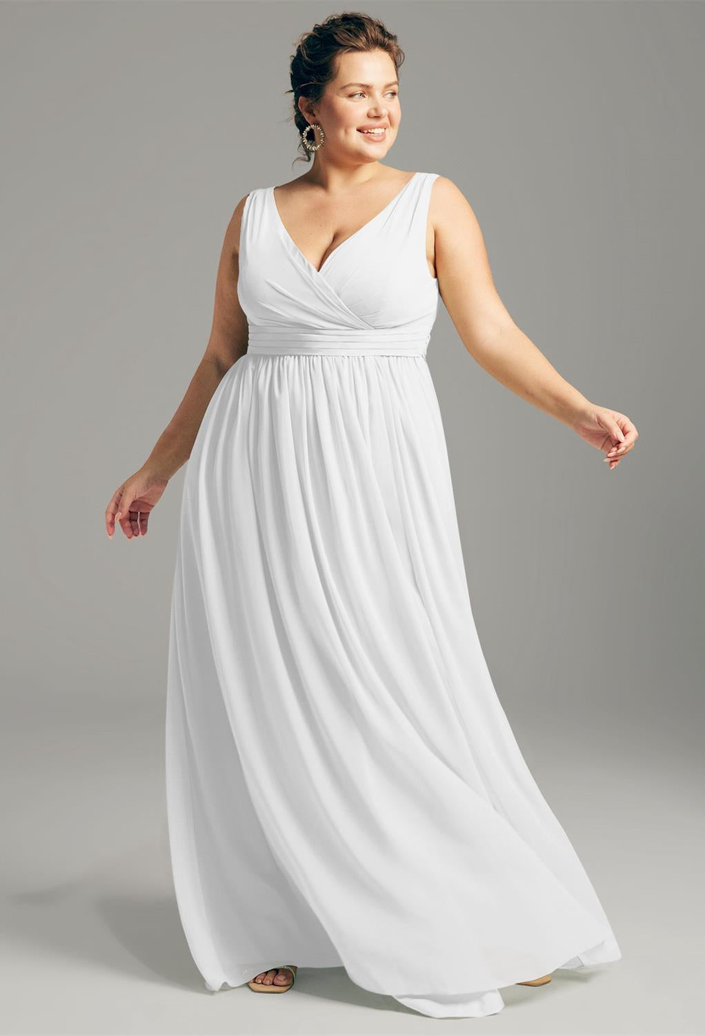 A woman wearing the "Hilaria" chiffon bridesmaid dress from Bergamot Bridal, a bridal shop in London, Ontario, stands against a plain background, looking to her left and smiling.