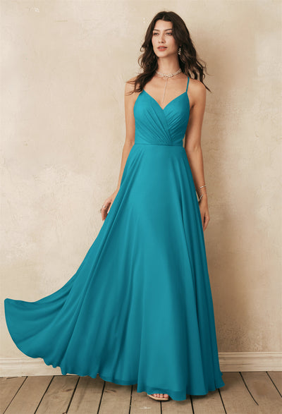 A woman in a teal, sleeveless evening gown stands gracefully against a textured wall on a wooden floor, radiating sophistication similar to the exquisite Melody chiffon bridesmaid dress from Bergamot Bridal's collection available in London, Ontario.