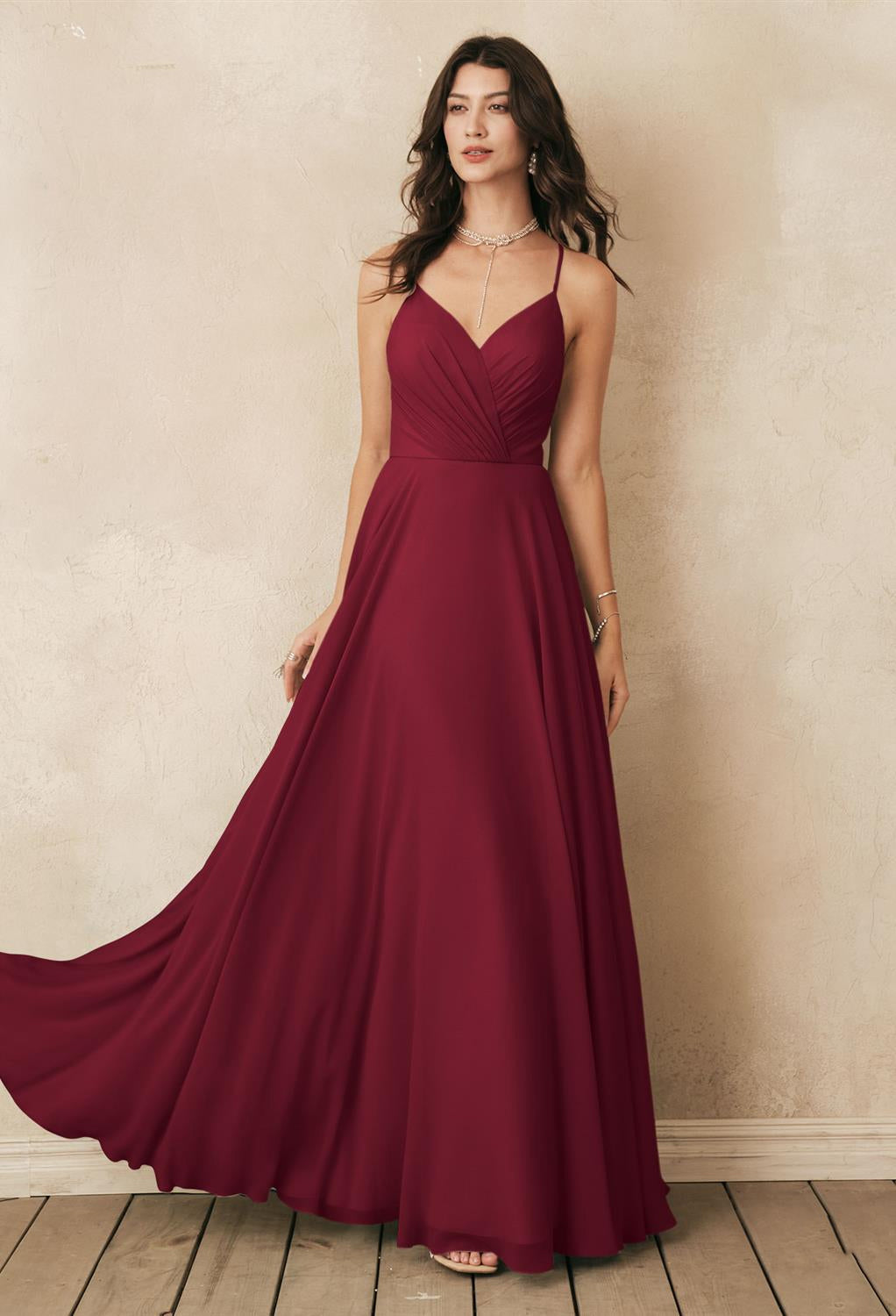 A woman wearing the "Melody" chiffon bridesmaid dress by Bergamot Bridal, suitable for any bridal shop in London, Ontario, stands against a textured cream wall. Her wavy hair and minimal jewelry perfectly complement the elegant look.