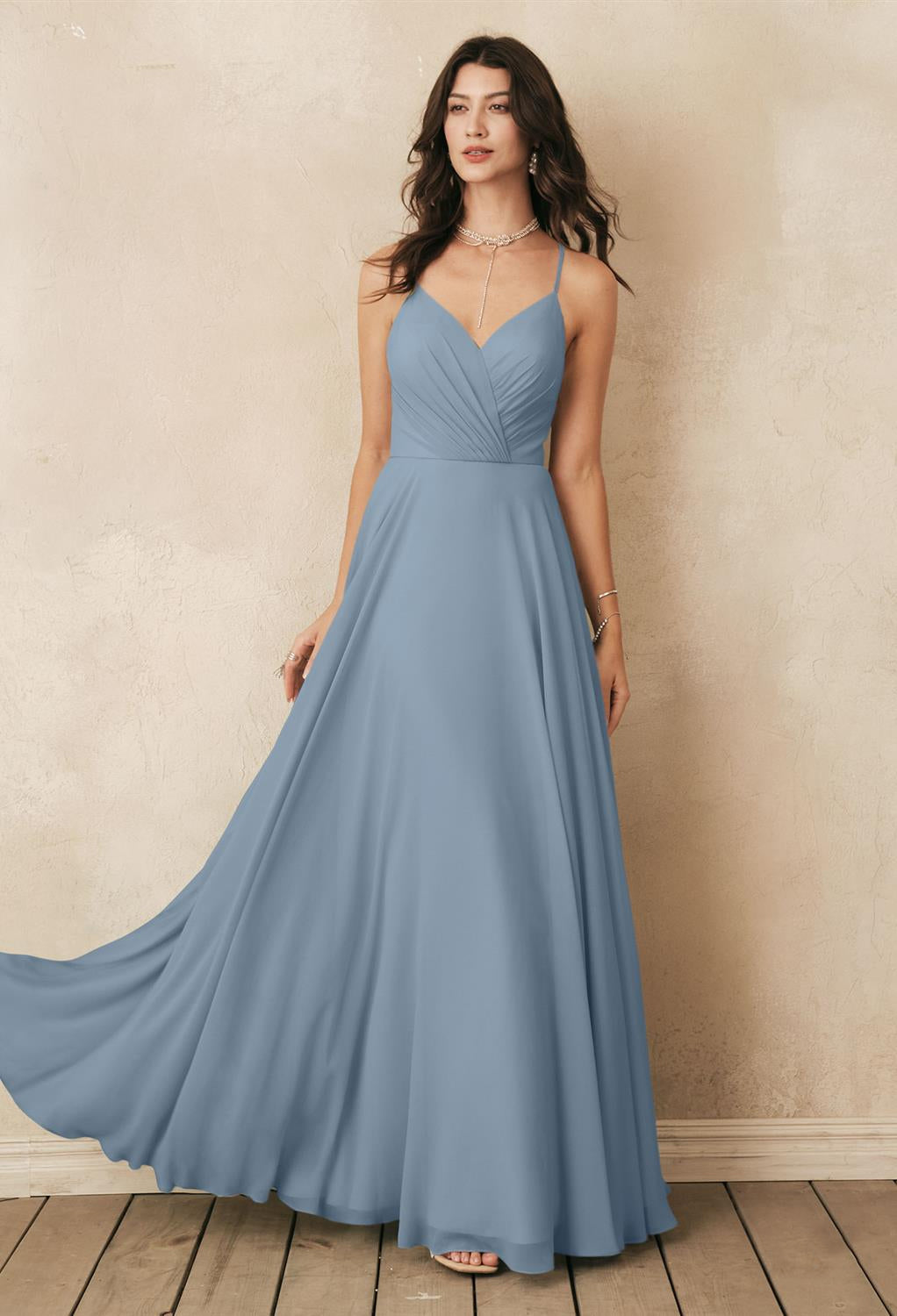 A person wearing a long, light blue dress with thin straps stands on a wooden floor against a textured beige wall, showcasing the affordable "Melody" chiffon bridesmaid dress from Bergamot Bridal, available at the bridal shop in London, Ontario.