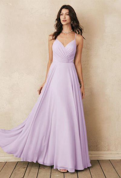 A woman in the Melody - Chiffon Bridesmaid Dress, designed by Bergamot Bridal, stands on a wooden floor against a textured wall, exuding elegance. This lilac gown is one of the many affordable wedding dress options available at our bridal shop in London, Ontario.