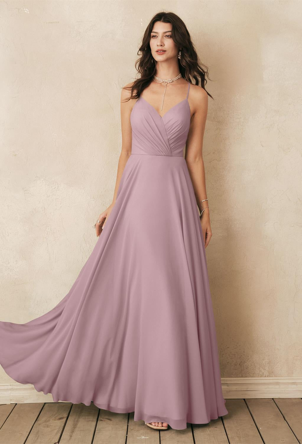 A woman in the Melody chiffon bridesmaid dress from Bergamot Bridal, a long, sleeveless mauve gown, stands against a textured beige wall on wooden flooring. She accessorizes with a necklace and bracelet, with wavy brown hair cascading over her shoulders, reminiscent of the affordable wedding dresses at bridal shops in London, Ontario.