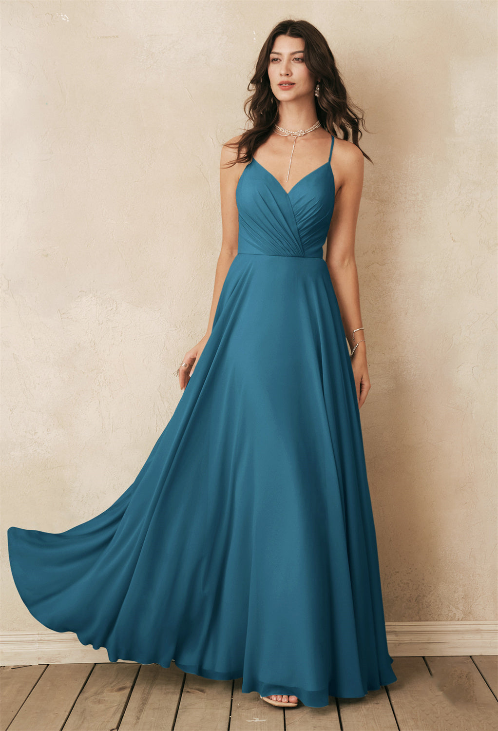 A woman wearing the Melody - Chiffon Bridesmaid Dress - Off the Rack by Bergamot Bridal, a teal sleeveless gown, stands on a wooden floor against a beige textured wall, reminiscent of the elegant selections found in bridal shops in London, Ontario.