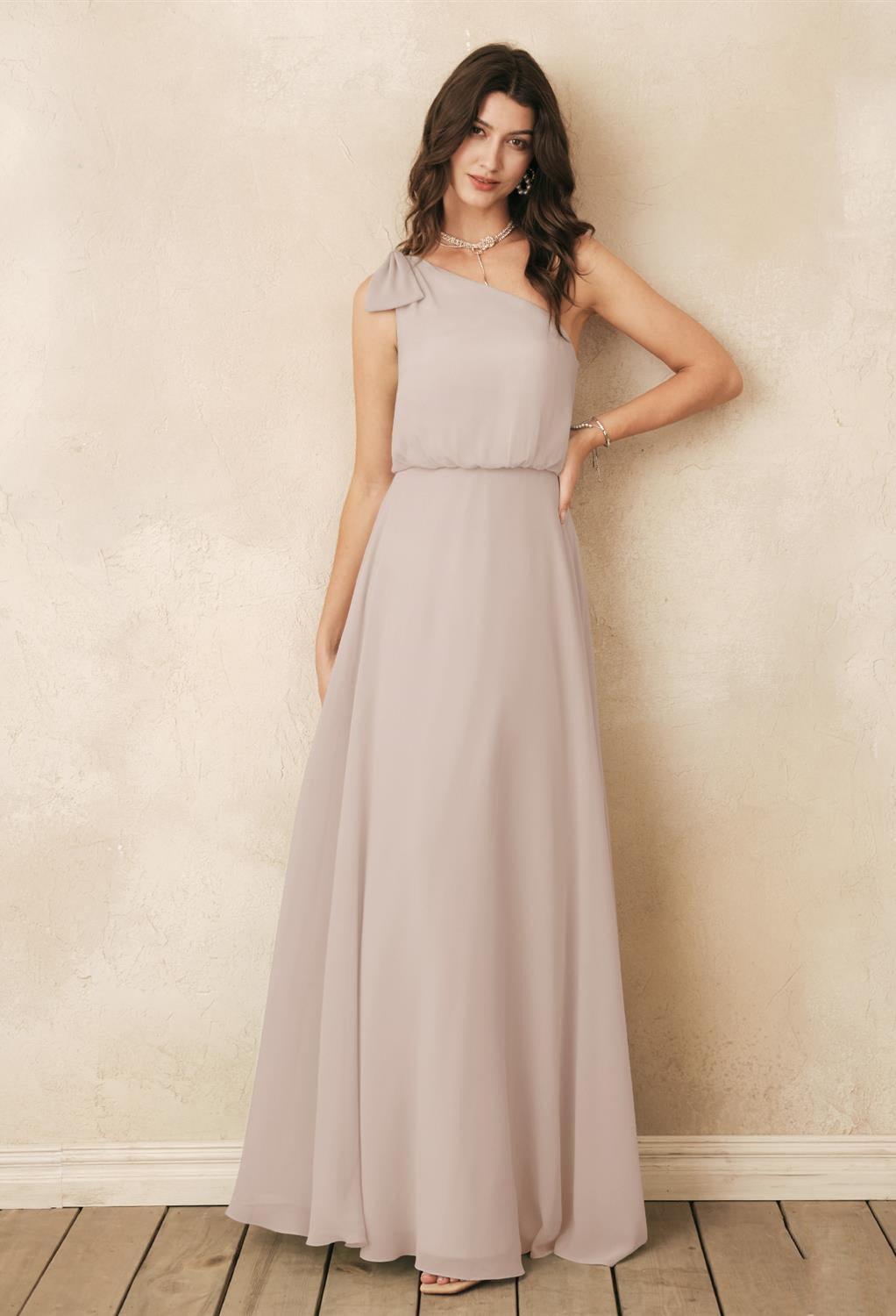 A woman in a sleeveless, light gray gown stands against a textured, cream-colored wall on a wooden floor, capturing the essence of elegance found in Bergamot Bridal's affordable Robina Chiffon Bridesmaid Dress - Off The Rack collection from London, Ontario.