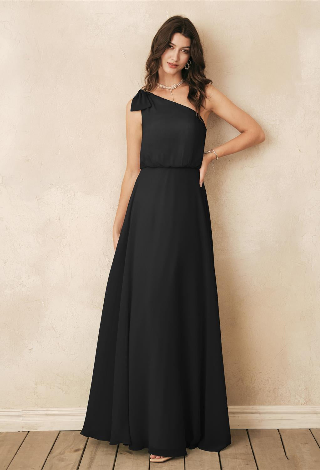 A woman dressed in the Robina Chiffon Bridesmaid Dress by Bergamot Bridal, with its sophisticated one-shoulder design in black, stands against a light textured backdrop on a wooden floor, capturing the classic elegance typically seen at bridal shops in London, Ontario.