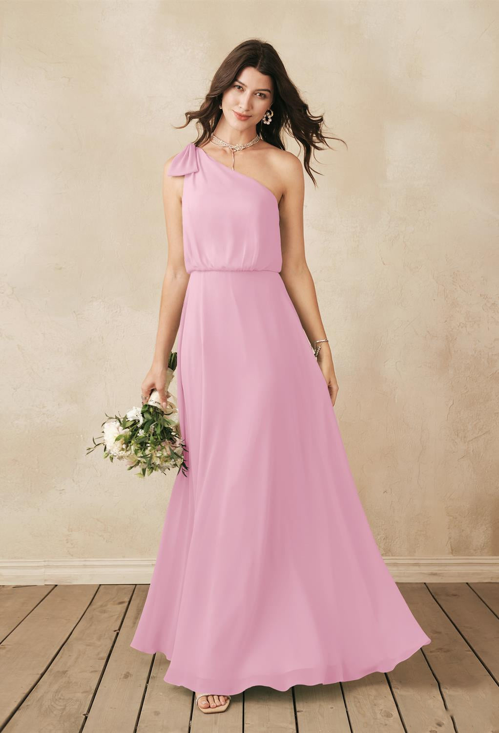 A woman in the Robina chiffon bridesmaid dress from Bergamot Bridal holds a bouquet of white flowers, standing on wooden flooring with a neutral textured background, capturing the elegance often seen in bridal shops London offers.