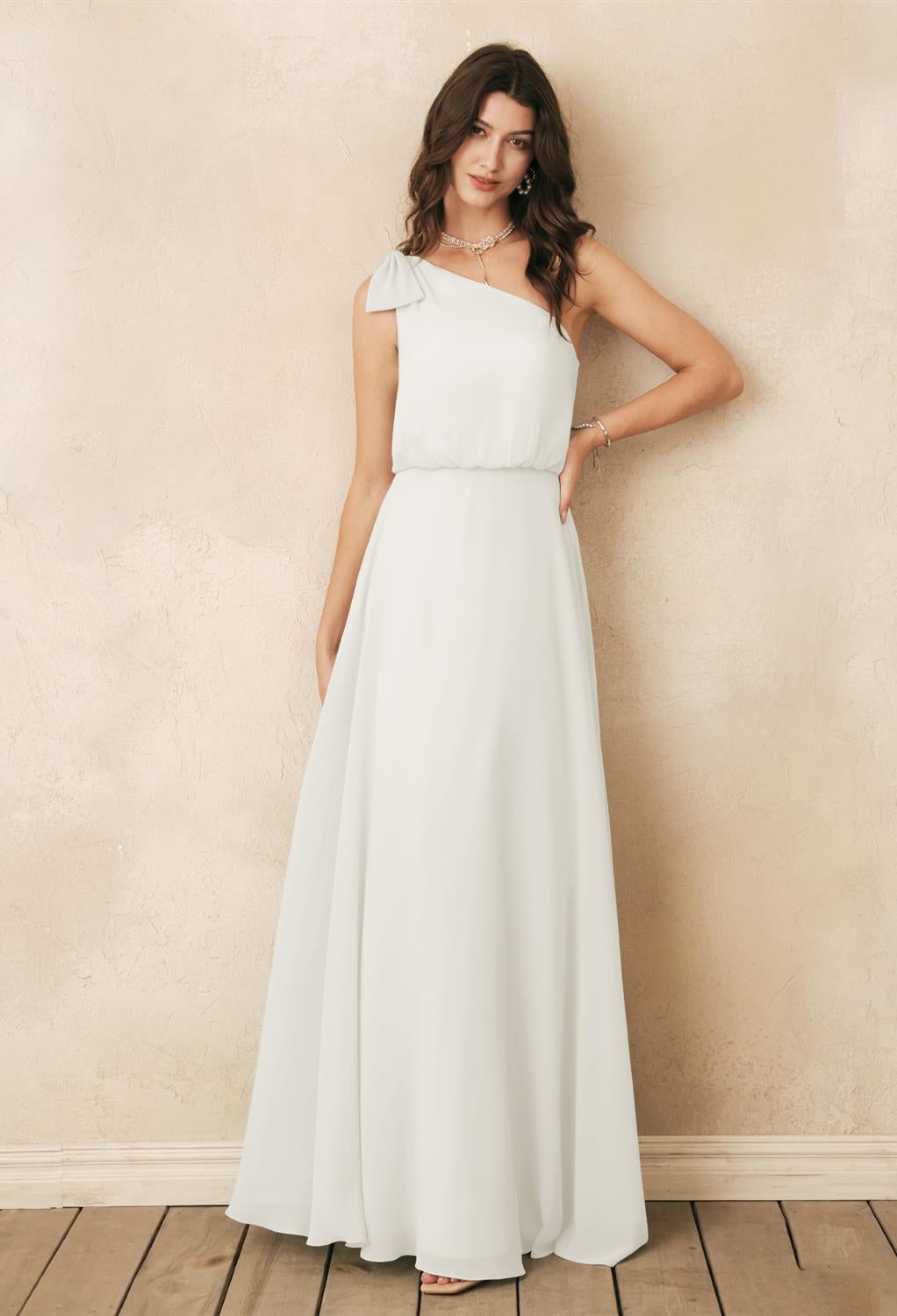 A woman wearing the Bergamot Bridal "Robina" - an off-the-rack chiffon bridesmaid dress, poses gracefully against a textured beige wall on a wooden floor, embodying the elegance found in London, Ontario's bridal shops.