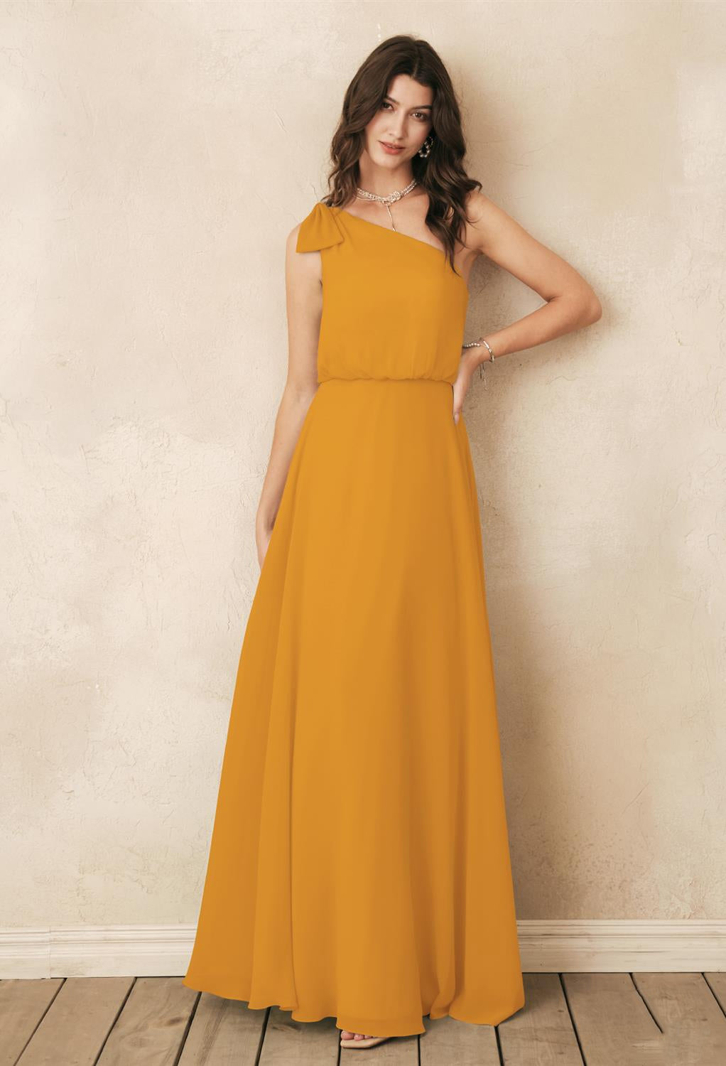 In a mustard yellow, one-shoulder gown, the woman stands gracefully against a textured beige wall on a wooden floor, embodying the elegance of the Robina Chiffon Bridesmaid Dress by Bergamot Bridal, reminiscent of those found in bridal shops across London.