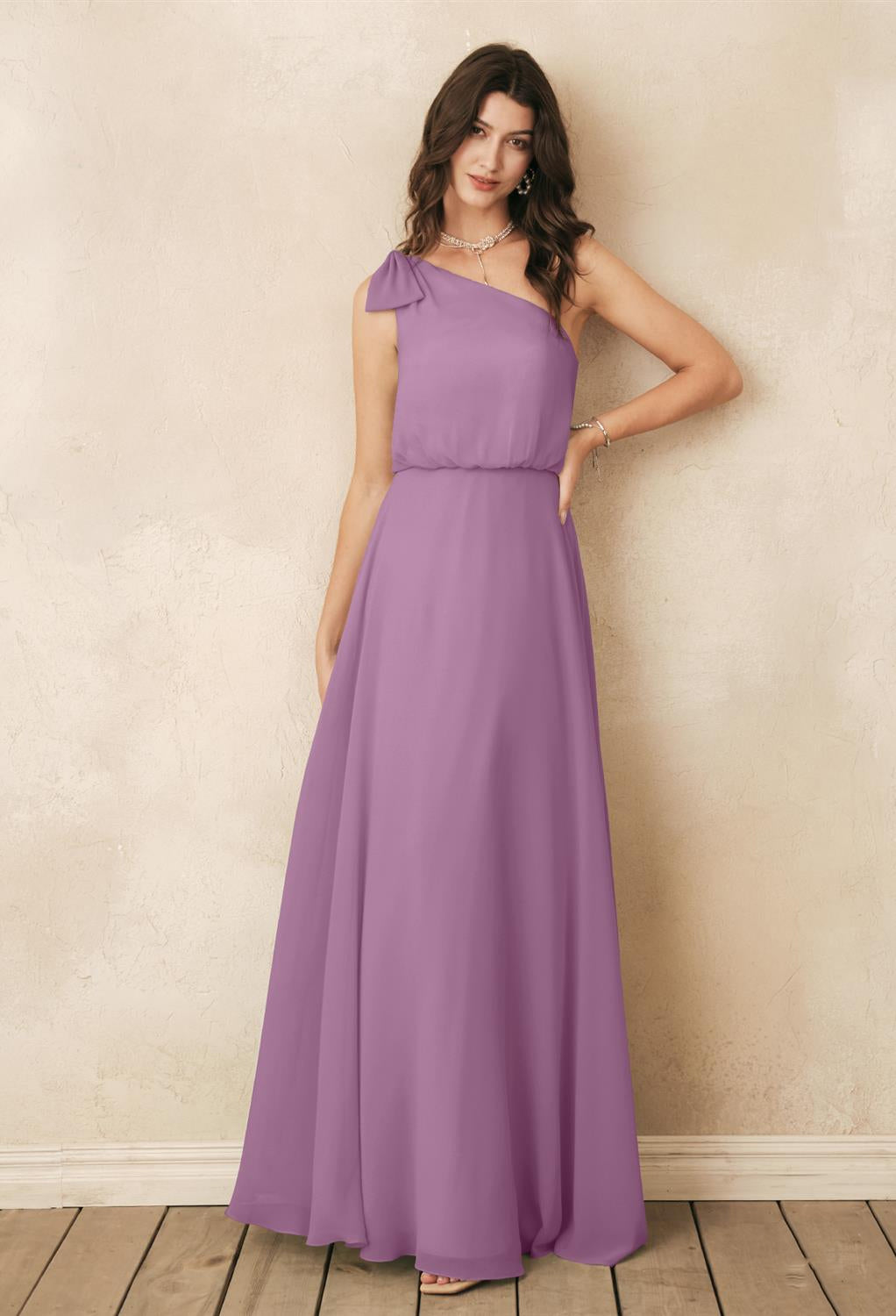 A woman in a Robina chiffon bridesmaid dress by Bergamot Bridal is positioned against a light-colored textured wall on a wooden floor, evoking the elegant options found in bridal shops in London, Ontario.