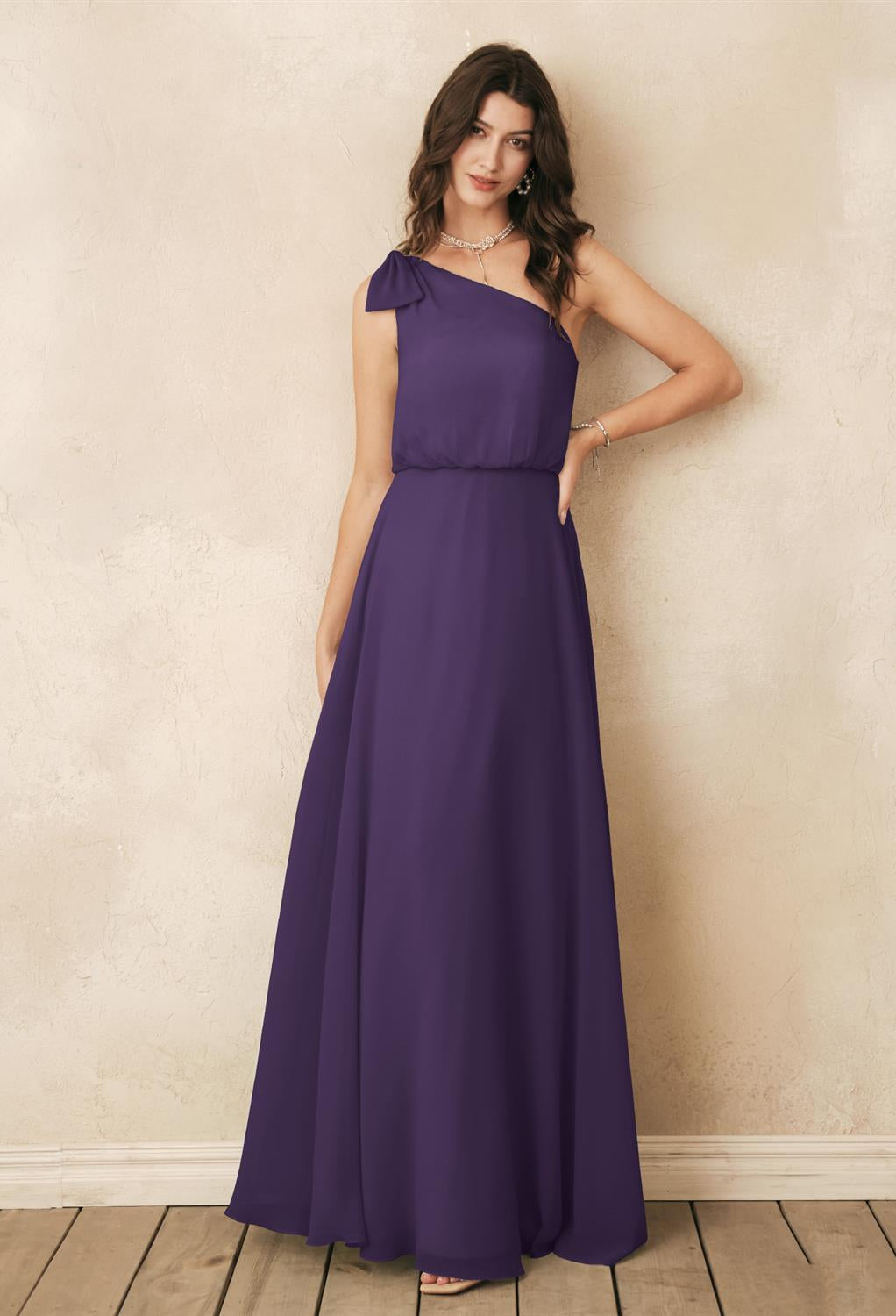 A woman in a one-shoulder, floor-length purple chiffon dress from Bergamot Bridal's Robina collection poses gracefully against a textured, light-colored wall, embodying elegance akin to the off-the-rack offerings at bridal shops in London, Ontario.