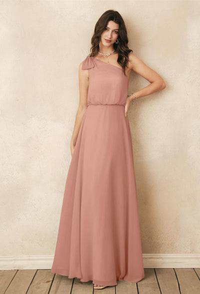 A woman wearing the Robina - Chiffon Bridesmaid Dress by Bergamot Bridal poses against a textured beige wall on wooden flooring, capturing the chic elegance of browsing bridesmaid dresses off the rack at a bridal shop in London, Ontario.