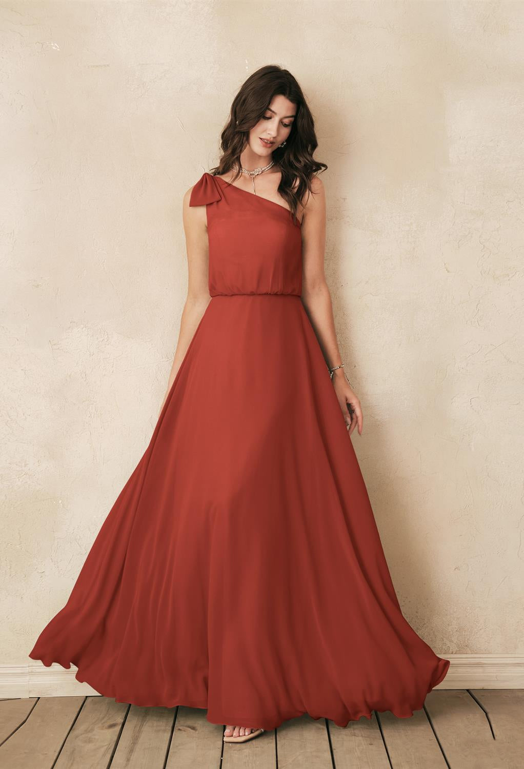 A woman in a long red dress, the Robina - Chiffon Bridesmaid Dress - Off The Rack by Bergamot Bridal, posing against a wall.