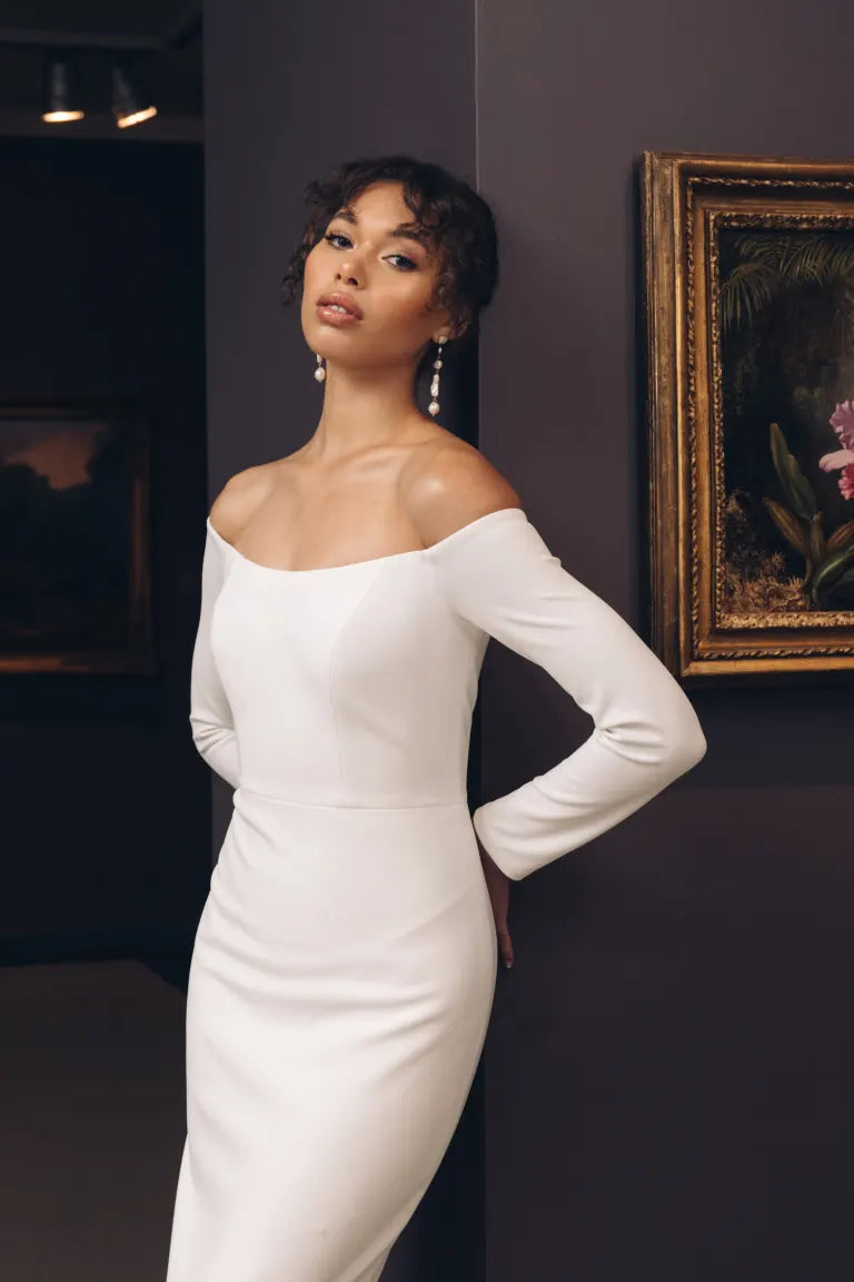 A woman in a white off the shoulder dress, the Ayla - Jenny Yoo Little White Dress from Bergamot Bridal, posing in front of paintings at a bridal shop in London.
