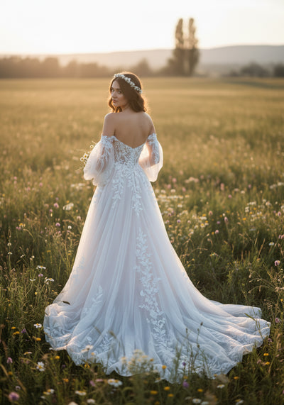 A woman in the Bergamot Bridal Floral Tulle A-line Gown with Off The Shoulder Sleeves stands in a grassy field at sunset, facing away and looking over her shoulder.