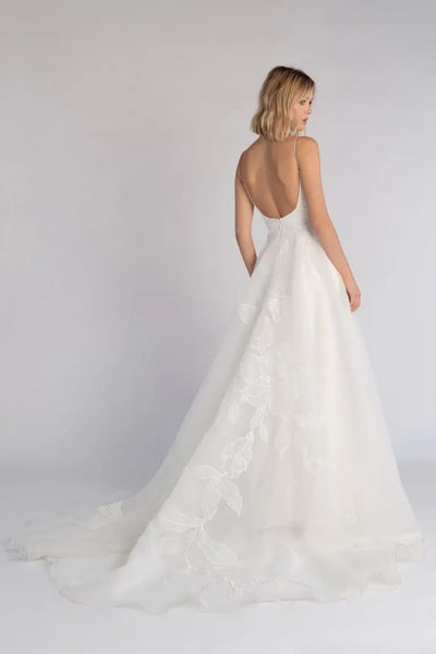 A woman standing with her back toward the camera, wearing an elegant white Abernathy - Jenny Yoo bridal gown made of romantic organza, featuring a low-cut back and a flowing train from Bergamot Bridal.