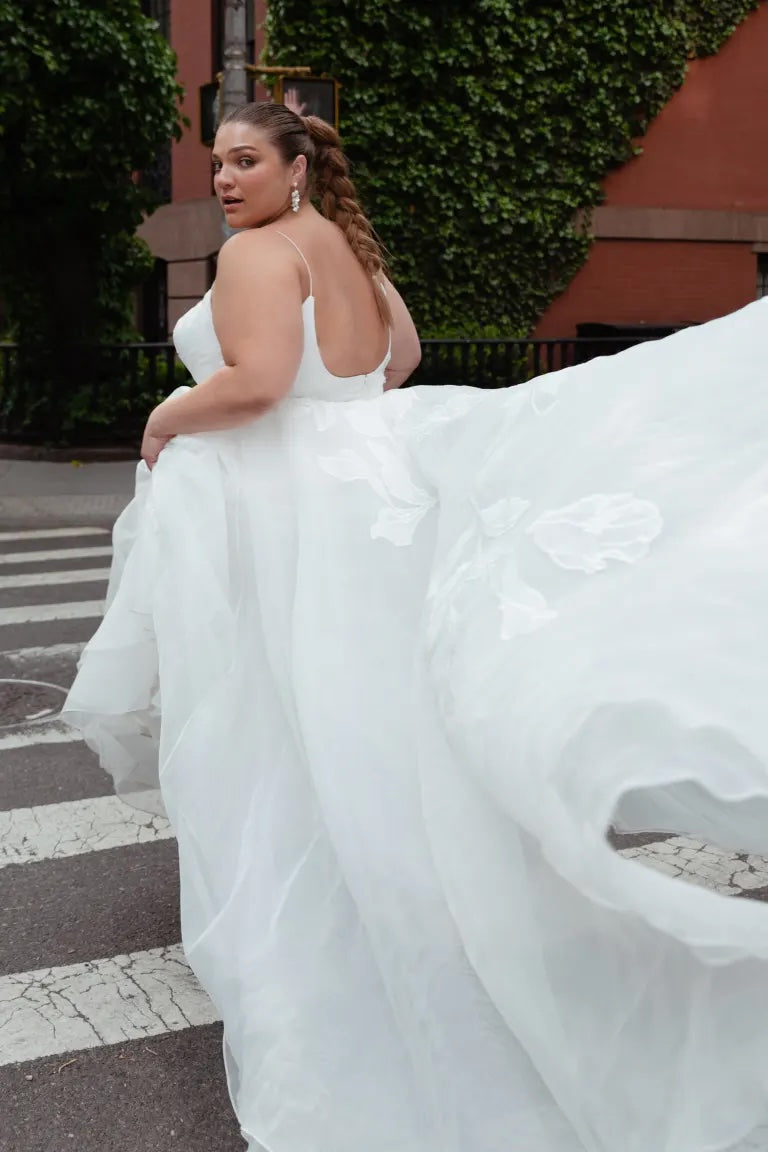A woman in an Abernathy - Jenny Yoo wedding dress with floral applique and a flowing train looks over her shoulder on a city street from Bergamot Bridal.