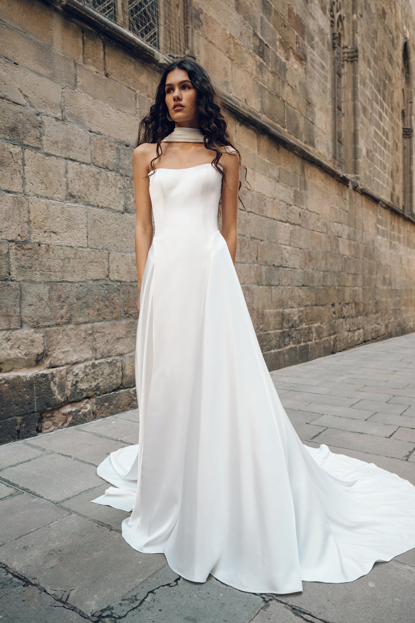 A woman wearing the Addilyn wedding dress by Bergamot Bridal stands on a stone street, leaning against a textured brick wall. The luxurious satin fabric flows elegantly with a subtle drop waist silhouette.