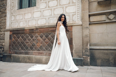 A woman in an elegant Bergamot Bridal Addilyn gown, designed by Jenny Yoo and crafted from luxurious satin, stands gracefully against a patterned stone wall.