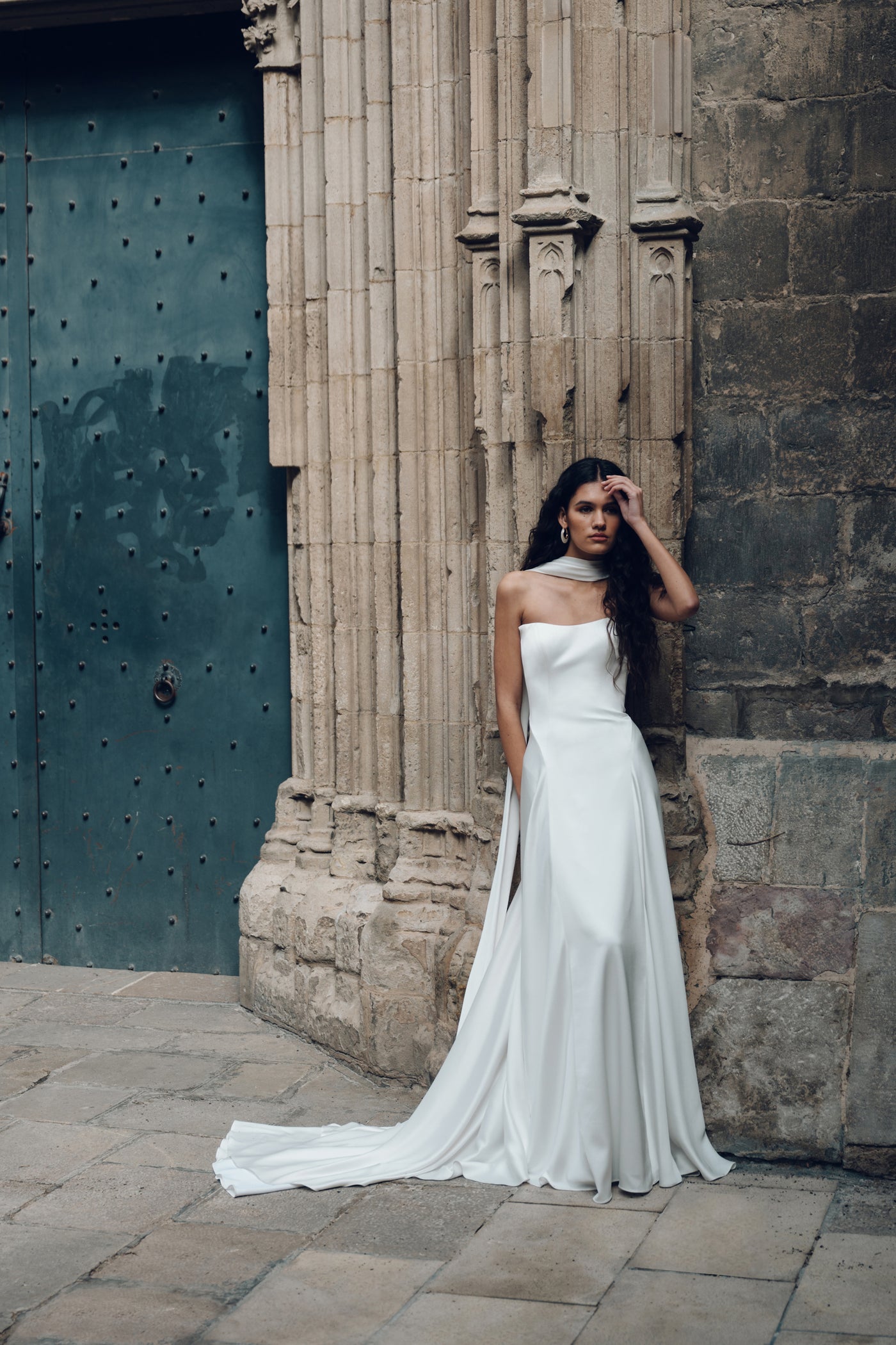 A woman in an Addilyn gown by Bergamot Bridal, crafted from luxe satin, stands against an ornate stone wall, with a large metal door nearby.