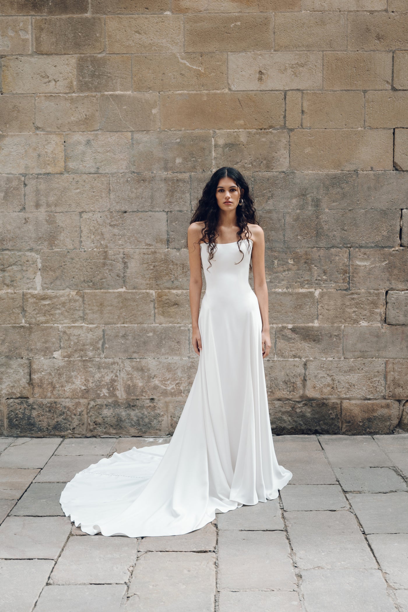 The woman, elegantly dressed in the Addilyn wedding dress by Bergamot Bridal, featuring a drop waist silhouette, stands gracefully on a stone pavement in front of a textured stone wall.