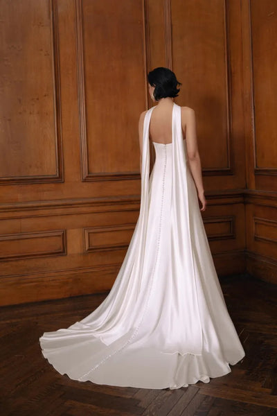 A woman in an elegant Jenny Yoo Wedding Dress gown made of luxe satin fabric with a trailing skirt stands with her back to the camera in a room with wooden paneling, exuding old Hollywood inspiration.