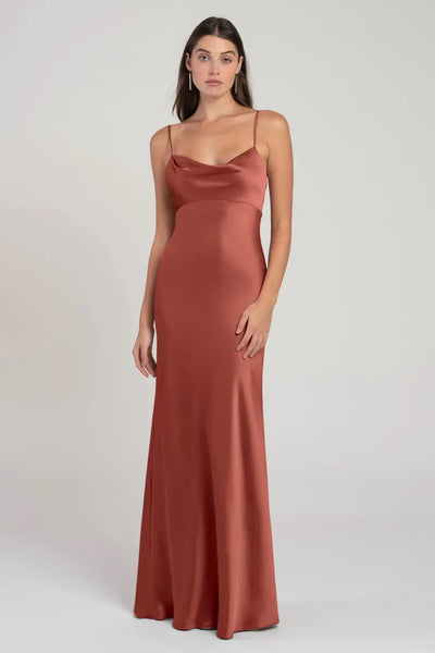 A woman wearing a long, satin rust-colored Addison bridesmaid dress by Jenny Yoo with spaghetti straps and an empire waist stands against a neutral background from Bergamot Bridal.