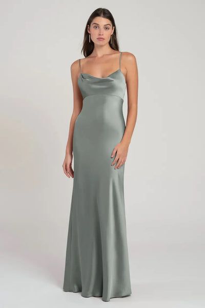 A woman standing against a neutral background, wearing a sleek, satin slip dress in sage green with spaghetti straps and an empire waist. The dress is the Addison Bridesmaid Dress by Jenny Yoo from Bergamot Bridal.