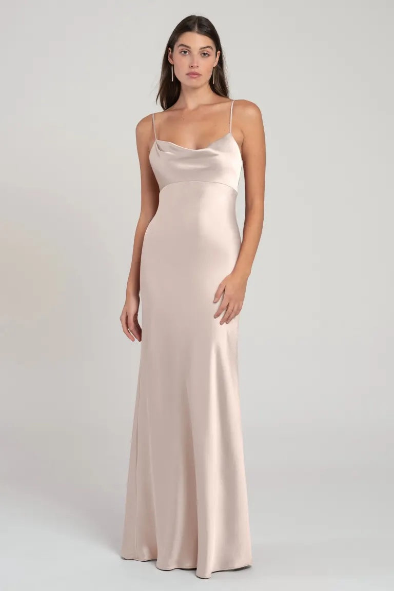 A woman wearing a long, sleeveless satin gown with an empire waist and satin cowl neck in a light neutral color, Addison - Bridesmaid Dress by Jenny Yoo, standing against a plain background.