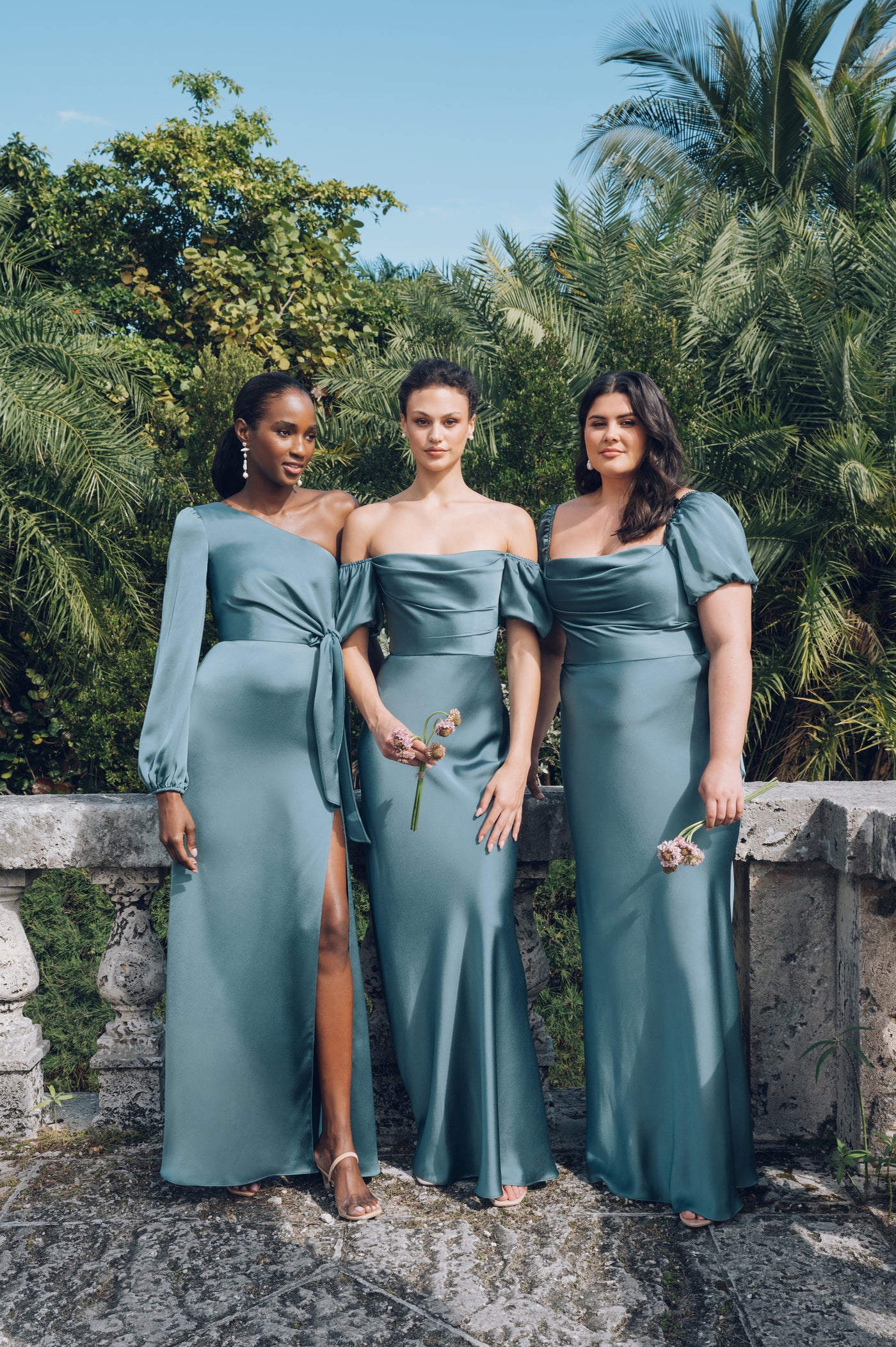 Three women wearing matching Jenny Yoo Bridesmaid Dresses with luxe satin fabric, standing together against a natural backdrop.
