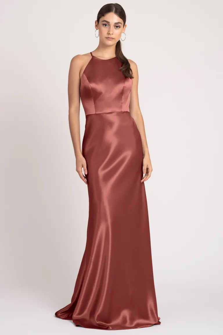 A woman in a sleek, satin back crepe, terracotta Alessia bridesmaid dress by Jenny Yoo with a high neckline stands posed against a neutral background.