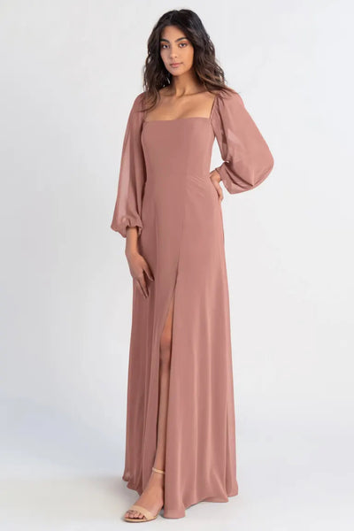 A woman posing in a long, dusty rose-colored chiffon Althea - Bridesmaid Dress by Jenny Yoo with a thigh-high slit and puff sleeves from Bergamot Bridal.