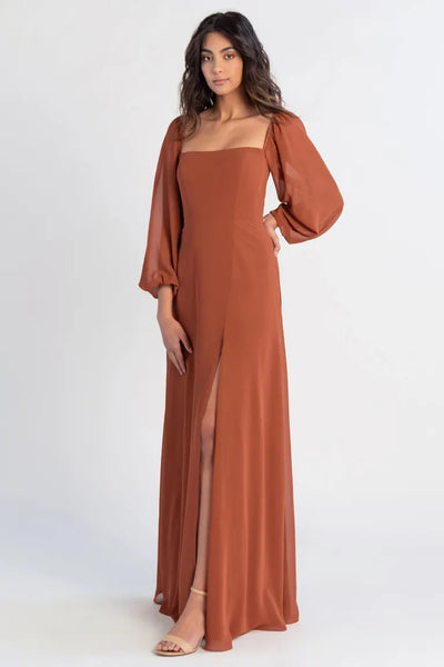 A woman poses in a sleek, long burnt orange Althea - Bridesmaid Dress by Jenny Yoo with a high leg slit and puff sleeves from Bergamot Bridal.