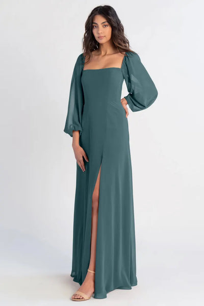 A woman in the Althea Bridesmaid Dress by Jenny Yoo Off The Rack from Bergamot Bridal, featuring a long teal chiffon design with puffed sleeves and a front slit, stands against a plain background, exuding bohemian beauty.