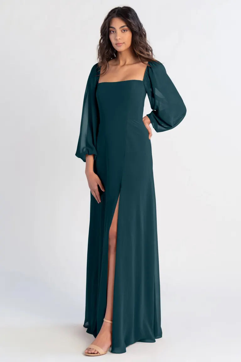 A woman in an Althea - Bridesmaid Dress by Jenny Yoo in a green chiffon evening gown with a high slit and balloon sleeves standing against a plain background. (Brand Name: Bergamot Bridal)
