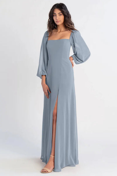 A woman models a blue chiffon Althea - Bridesmaid Dress by Jenny Yoo with a high slit and long sleeves from Bergamot Bridal.