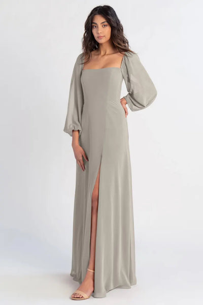 A woman poses in a long, flowing beige chiffon Althea bridesmaid dress by Jenny Yoo with a thigh-high slit and flowing sleeves from Bergamot Bridal.