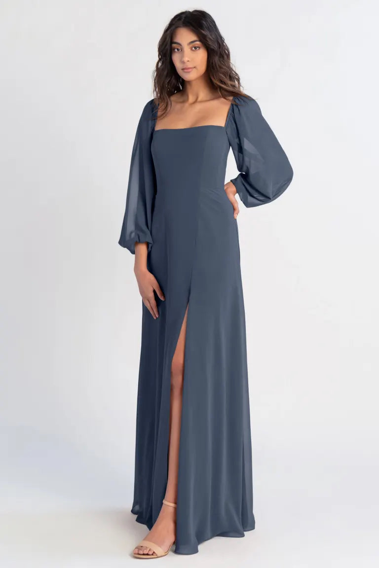 Woman modeling a long grey chiffon Althea bridesmaid dress by Jenny Yoo with a leg slit and flowing sleeves against a white background from Bergamot Bridal.
