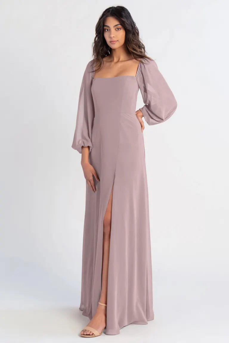 A woman modeling an elegant, long mauve Althea - Bridesmaid Dress by Jenny Yoo chiffon dress with a thigh-high slit and draped sleeves from Bergamot Bridal.