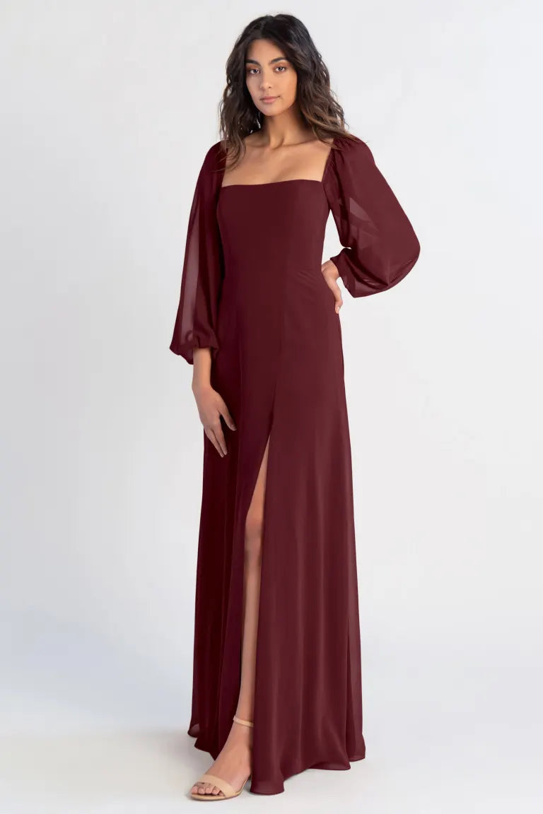 A woman posing in an elegant burgundy bridesmaid dress with a thigh-high slit and sheer long sleeves from the Althea - Bridesmaid Dress by Jenny Yoo from Bergamot Bridal.