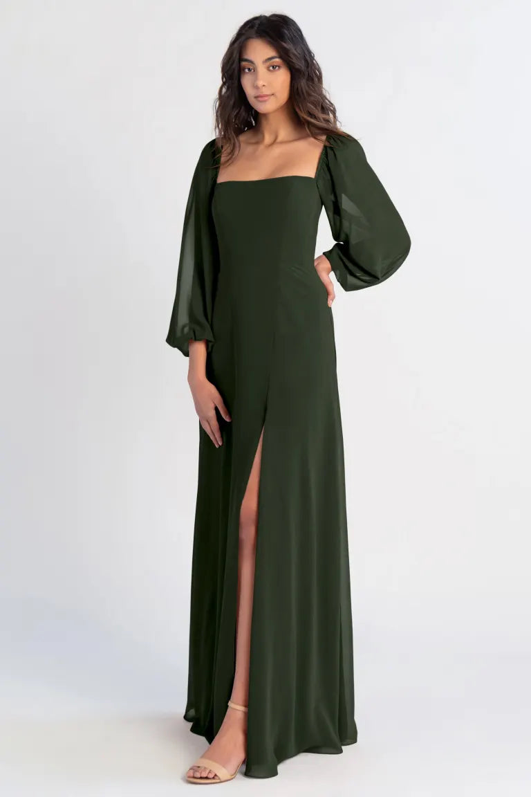 Clad in elegance, the woman poses gracefully in the Althea Bridesmaid Dress by Jenny Yoo Off The Rack from Bergamot Bridal, a dark green gown with long sleeves and a high slit set against a plain backdrop.