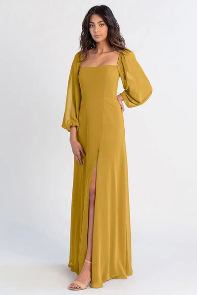 Woman posing in a long mustard chiffon evening dress with a thigh-high slit, the Althea - Bridesmaid Dress by Jenny Yoo from Bergamot Bridal.