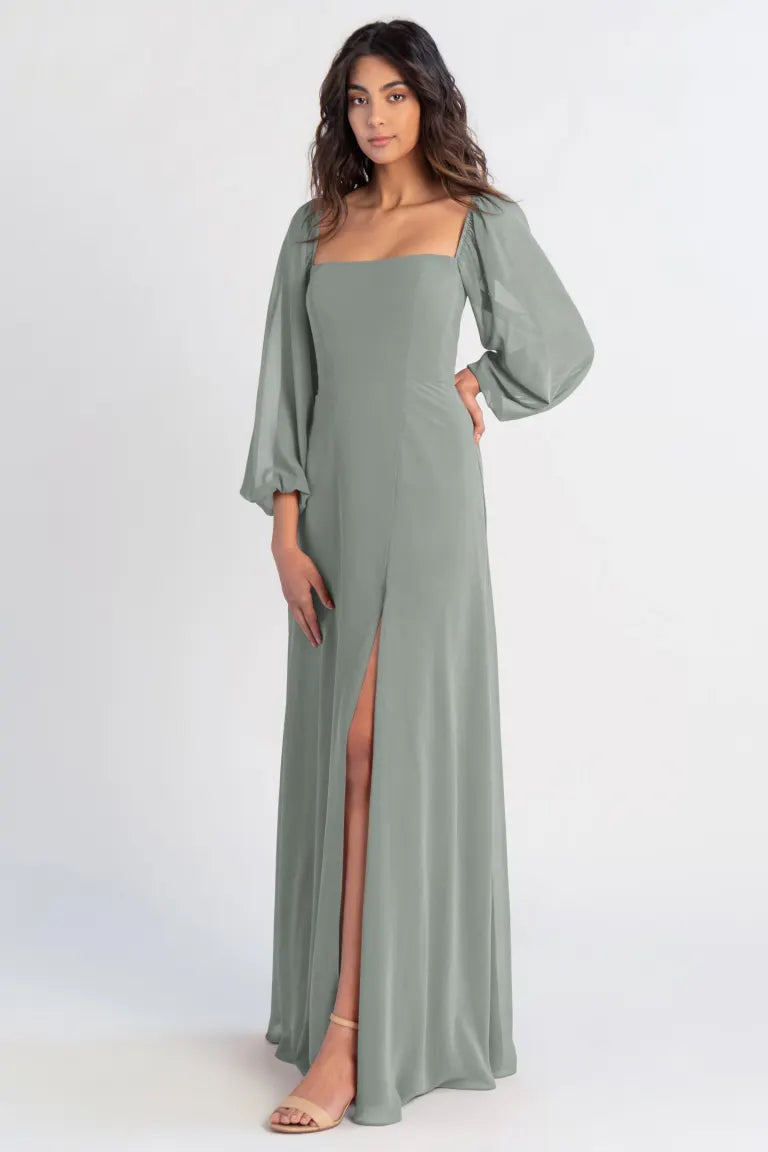 A woman posing in a Althea - Bridesmaid Dress by Jenny Yoo in sage green chiffon with a high slit and lantern sleeves from Bergamot Bridal.