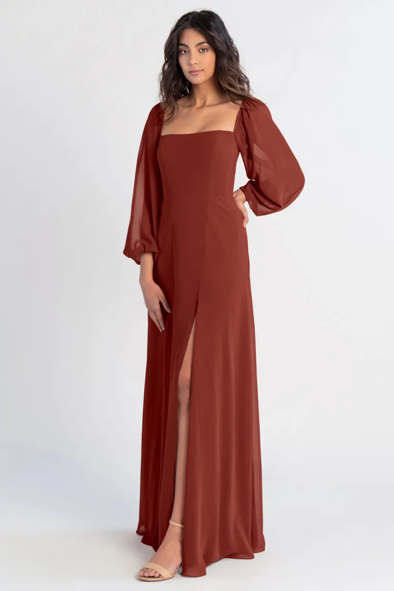 A woman posing in a sleek, rust-colored Althea - Bridesmaid Dress by Jenny Yoo with a thigh-high slit and billowy sleeves from Bergamot Bridal.