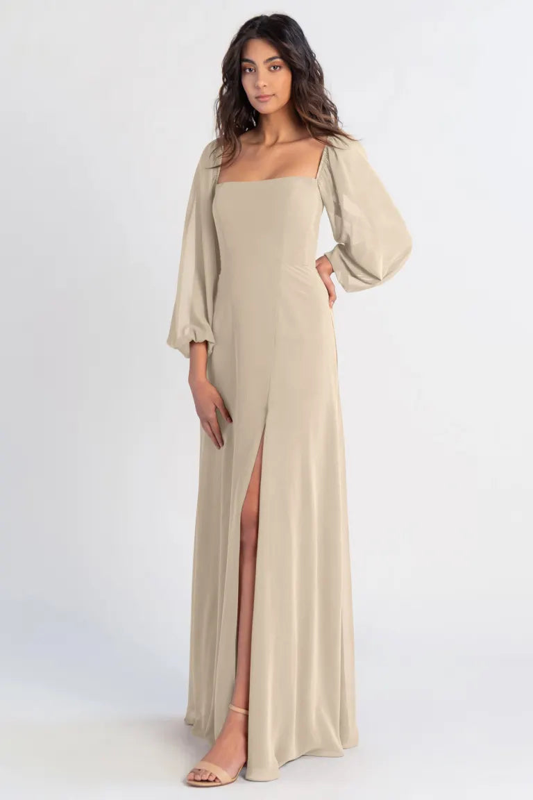 A woman posing in an elegant beige bridesmaid dress with a high slit and flowing chiffon sleeves, the Althea - Bridesmaid Dress by Jenny Yoo from Bergamot Bridal.