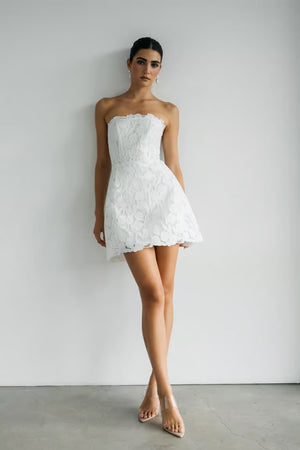 A woman poses against a white wall in the Amanda - Jenny Yoo Little White Dress by Bergamot Bridal, a strapless short lace dress ideal for bridal events, styled with clear heeled sandals.