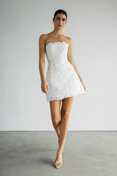 A woman stands indoors in the Amanda - Jenny Yoo Little White Dress by Bergamot Bridal, a strapless white lace mini dress perfect for bridal events, paired with clear high heels against a plain backdrop.