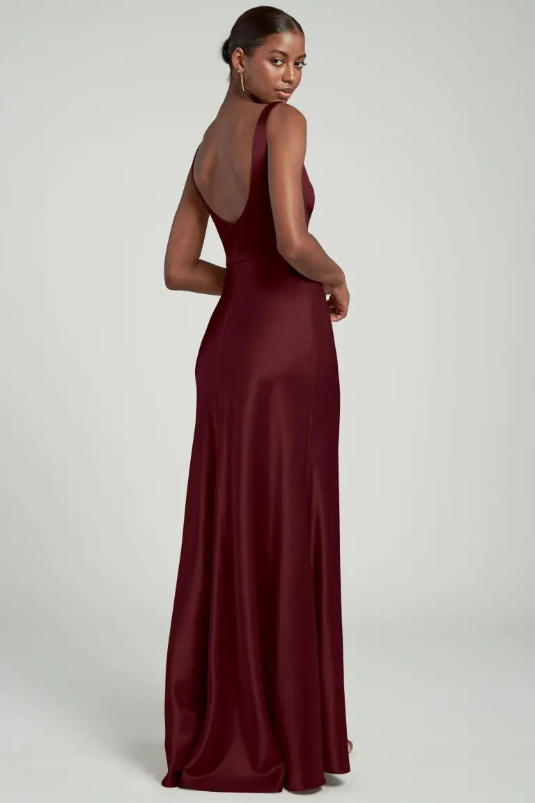 Woman wearing a sleek burgundy satin back crepe evening gown with a square neck, looking over her shoulder, the Ariana Bridesmaid Dress by Jenny Yoo from Bergamot Bridal.