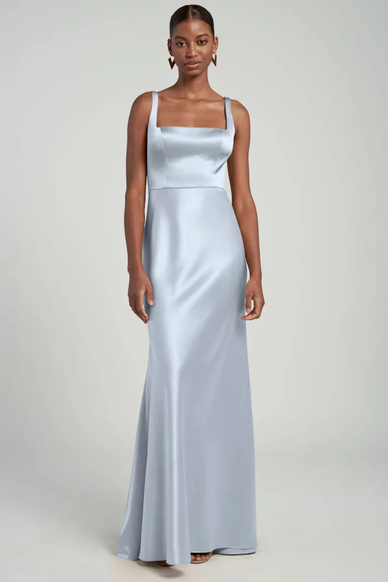 A person standing in a studio, wearing the elegant, floor-length "Ariana - Bridesmaid Dress" by Jenny Yoo from Bergamot Bridal. The dress is crafted from light blue satin and features a square neckline and thin straps, with a bias cut skirt that gracefully enhances its silhouette.