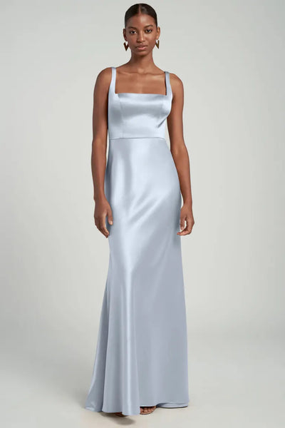 A person standing in a studio, wearing the elegant, floor-length "Ariana - Bridesmaid Dress" by Jenny Yoo from Bergamot Bridal. The dress is crafted from light blue satin and features a square neckline and thin straps, with a bias cut skirt that gracefully enhances its silhouette.